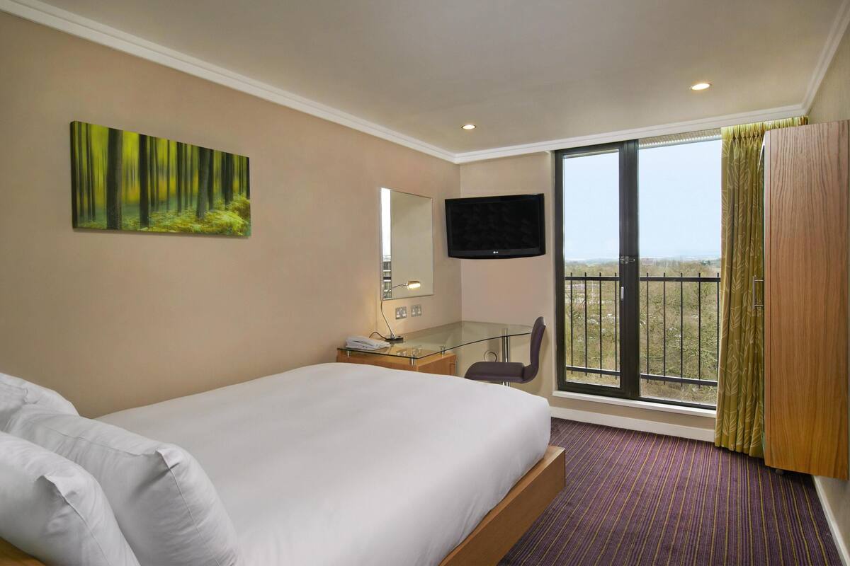 A hotel room with a bed, a desk with a telephone, a chair, a TV mounted on the wall, and a large window with a view.