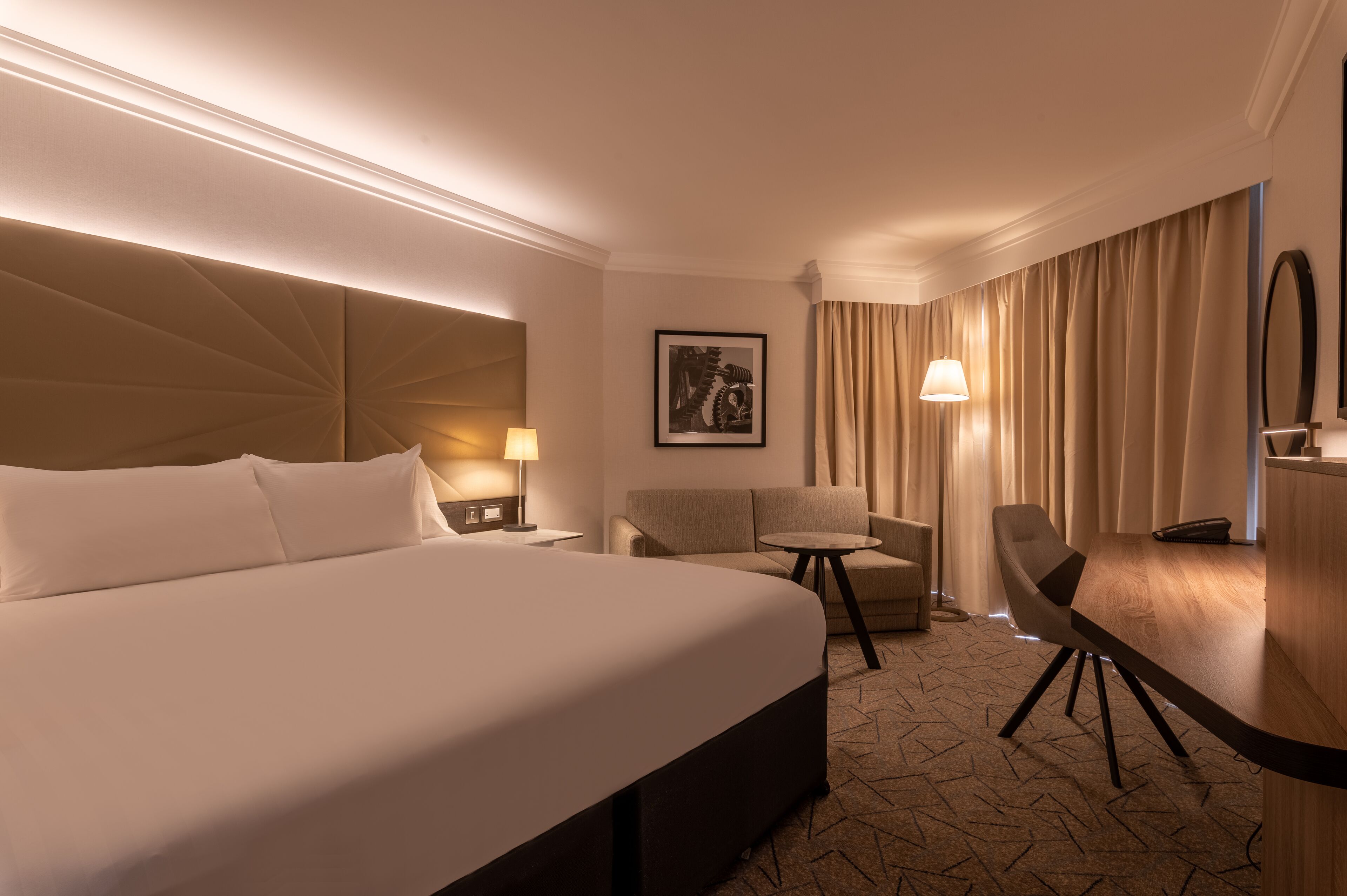 Ein Hotelzimmer mit einem großen Bett, einem Schreibtisch, einem Stuhl, einer Lampe und einem gerahmten Bild an der Wand.
