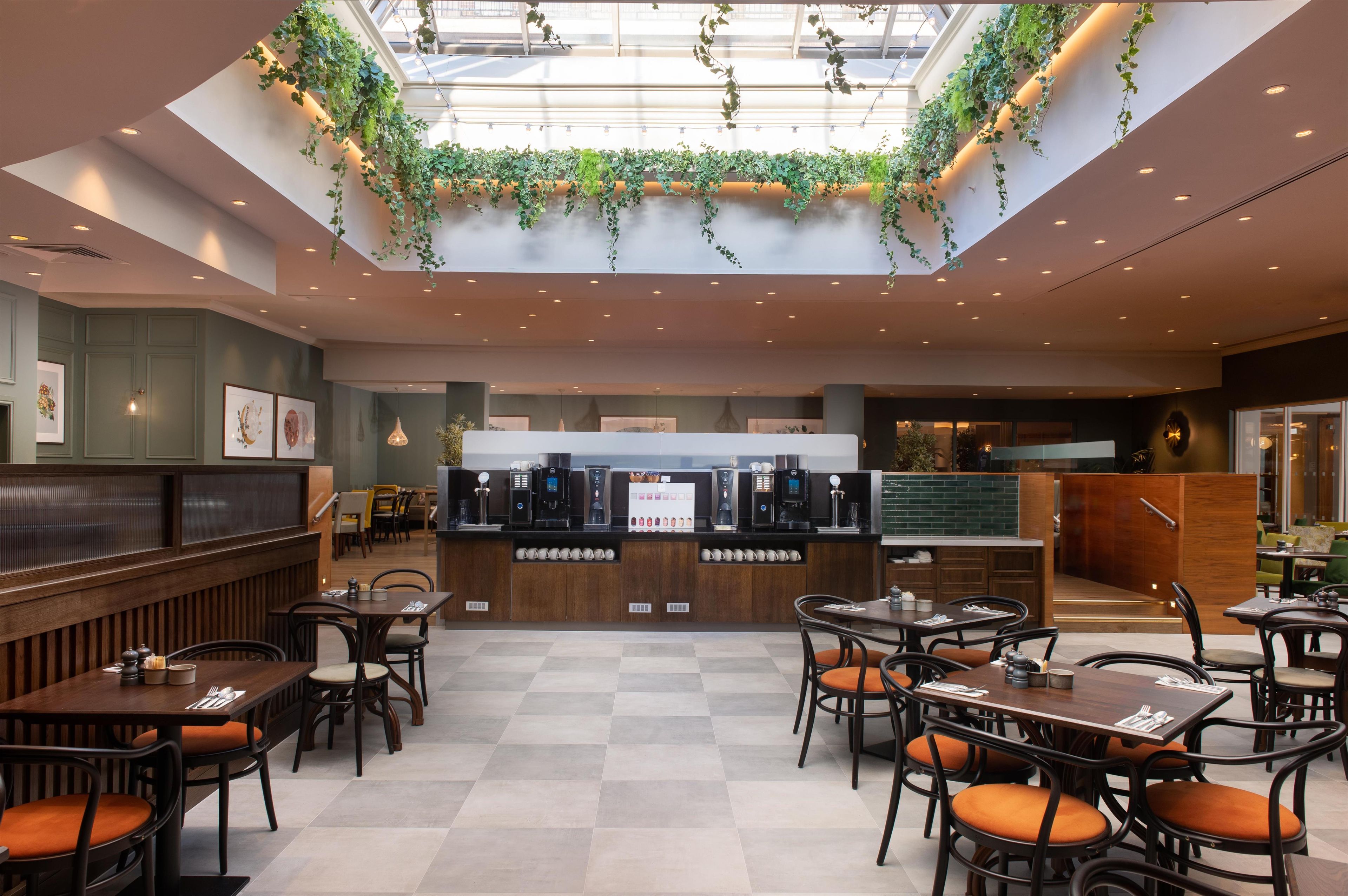 Una cafetería moderna con mostrador central, zona de estar y tragaluz con plantas.