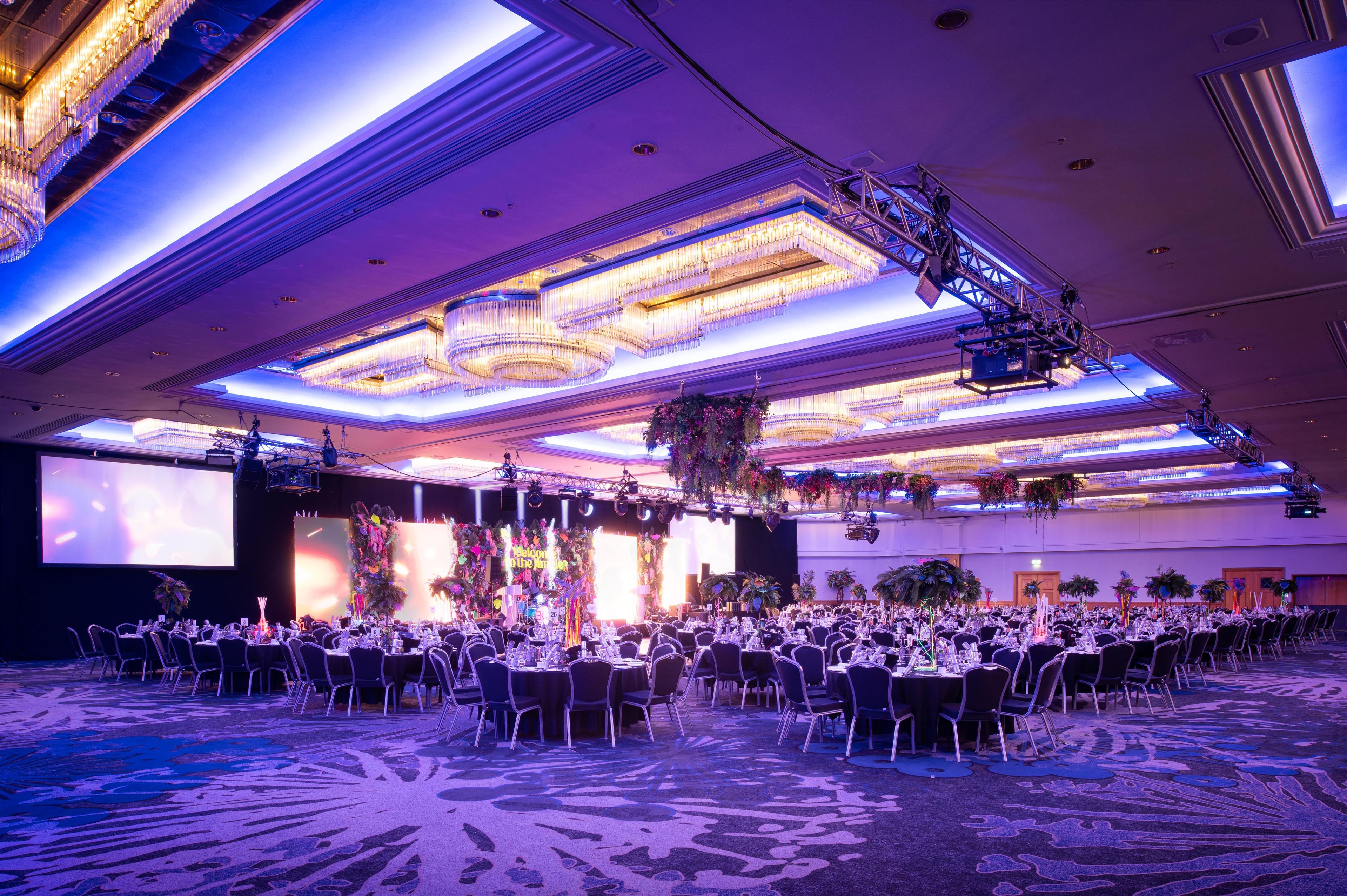 A large banquet hall with rows of tables and chairs, a stage with a backdrop, and a ceiling with chandeliers.