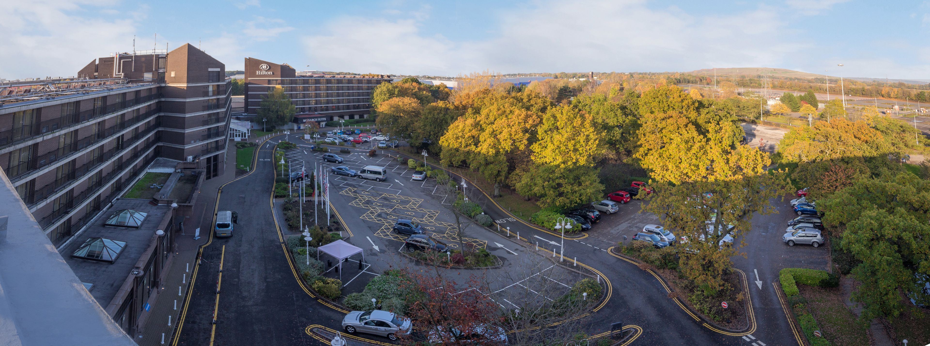 A multi-story hotel with a parking lot, a roundabout, and surrounding trees.