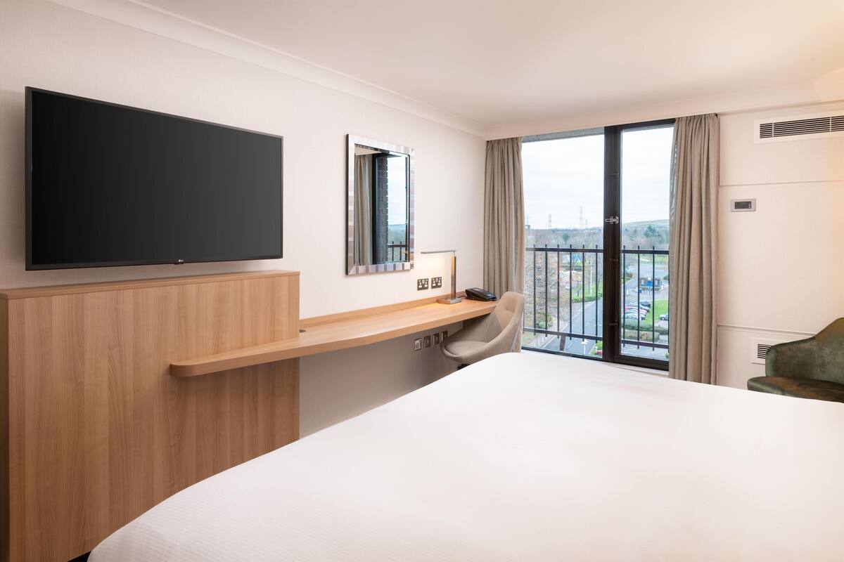 A hotel room with a large bed, a TV mounted on the wall, a desk with a chair, and a view of the city through the window.