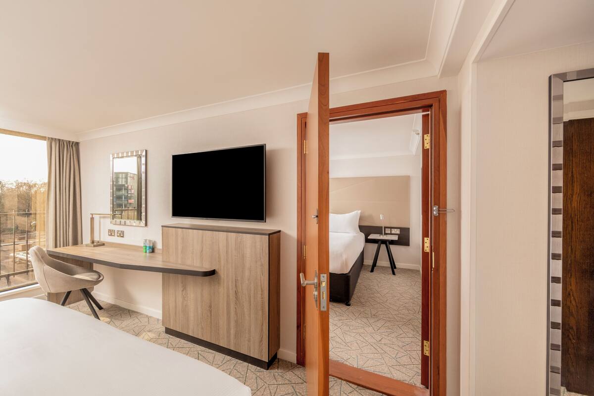 A hotel room with a bed, a desk with a chair, a television, a mirror, and a door leading to another room.