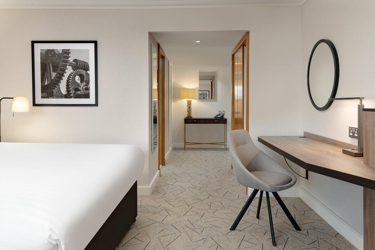 A hotel room with a bed, a desk with a chair, a desk lamp, a framed picture on the wall, and a patterned floor.