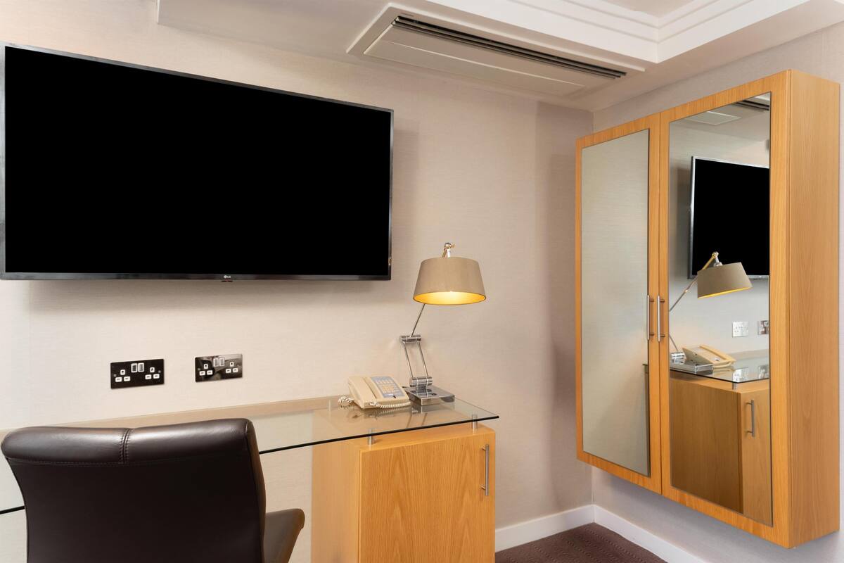 A hotel room with a flat-screen TV, a desk with a lamp, a telephone, and a wooden cabinet with a mirror.