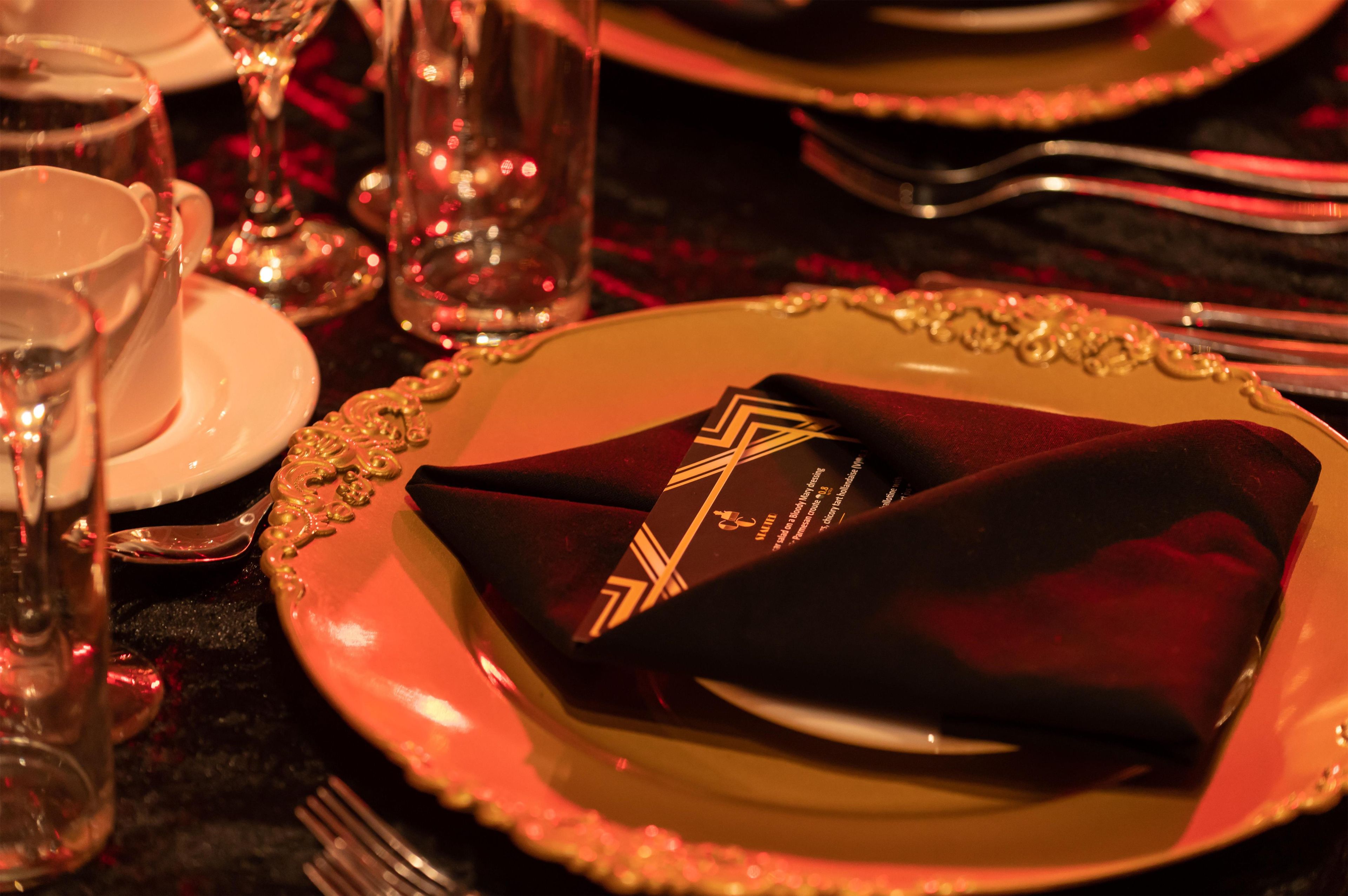A formal dining setting with a red napkin, a gold-rimmed plate, and a glass of water.