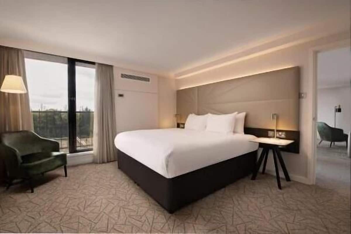 A hotel room with a large bed, a chair, a nightstand, and a view through a large window.