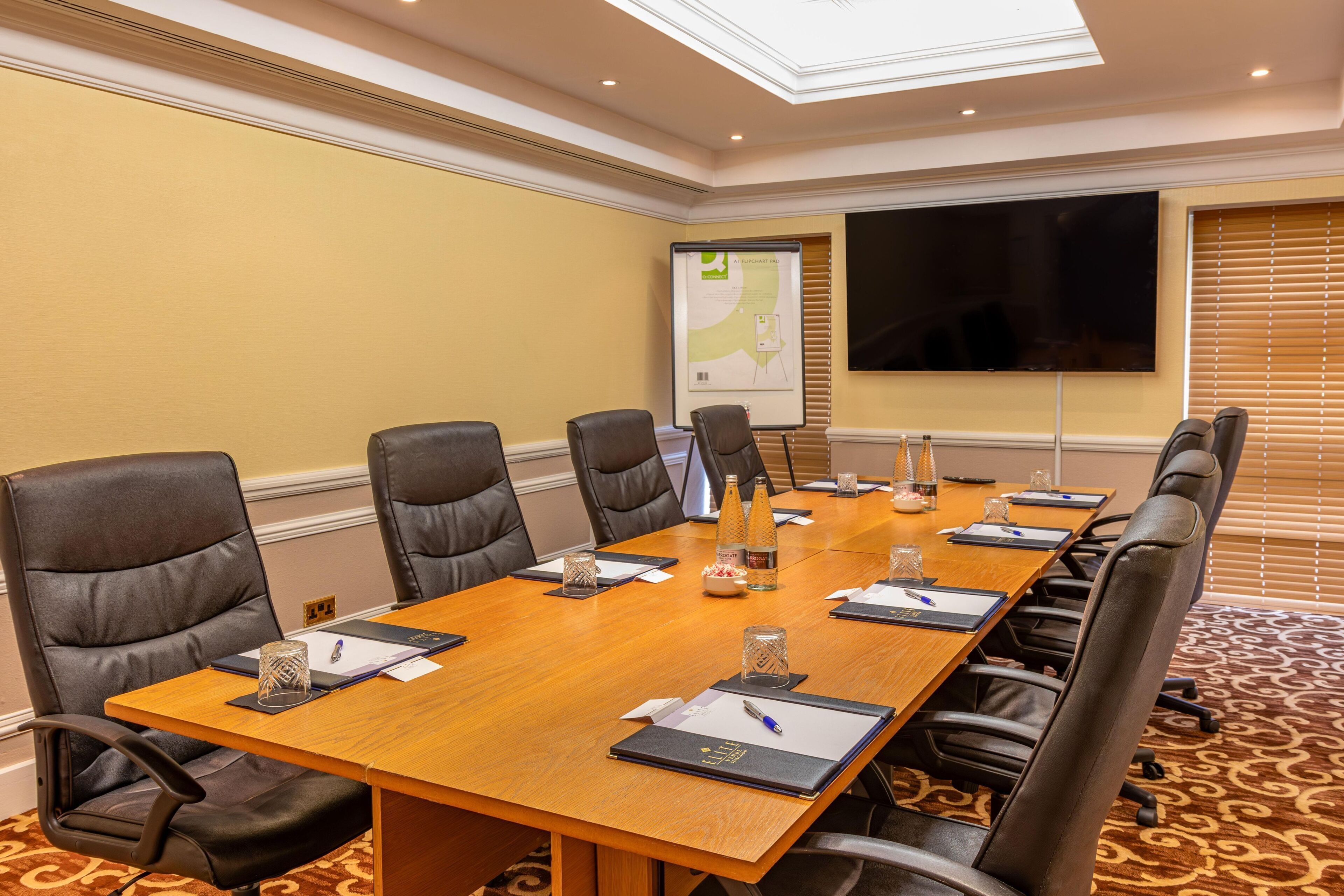 A conference room with a long table, black leather chairs, a projector screen, and a large TV.