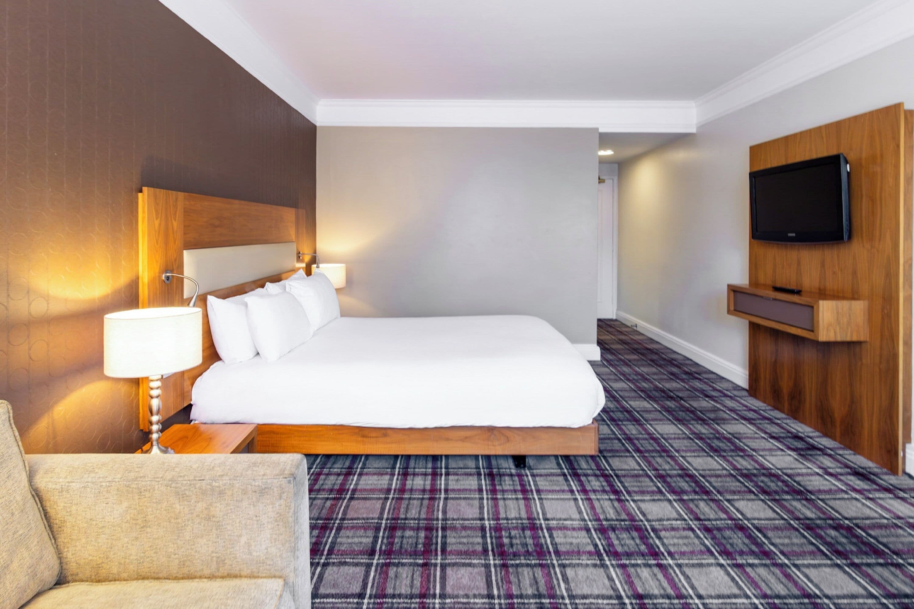 Ein Hotelzimmer mit einem großen Bett, einem an der Wand montierten Fernseher, einem karierten Teppich und einem hölzernen Kopfteil.