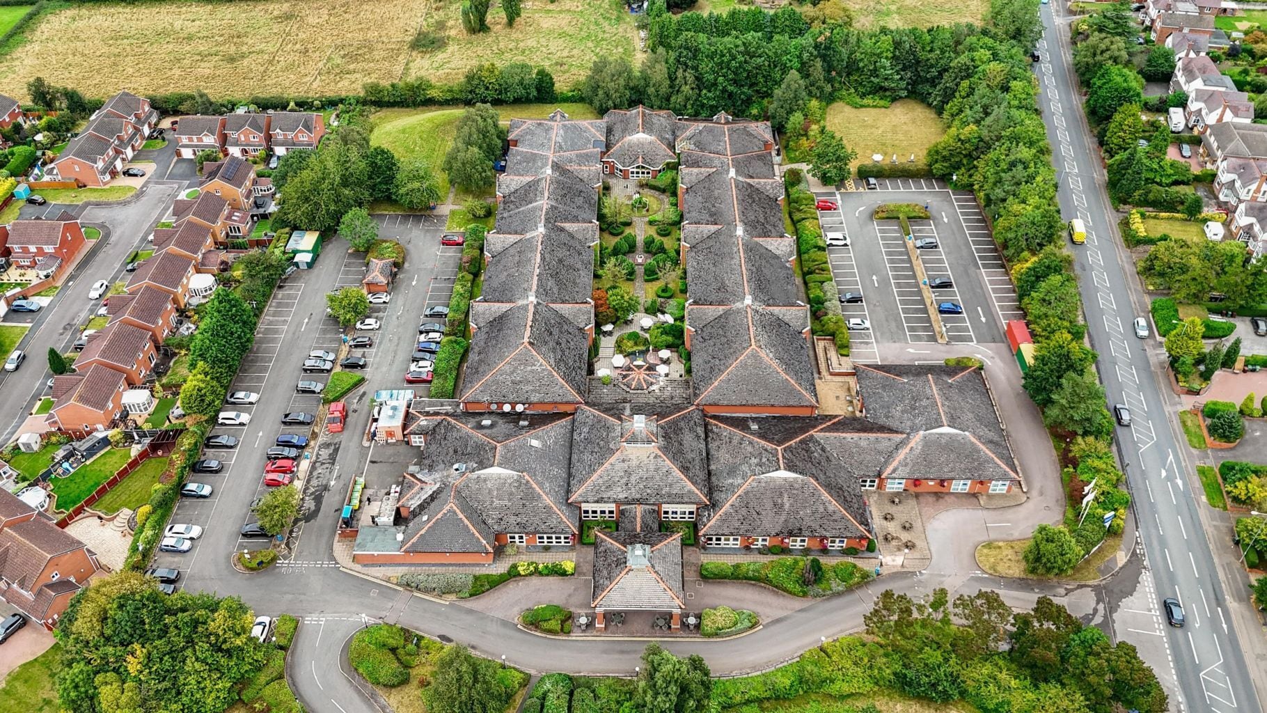 Vista aérea de una zona residencial con un edificio central que destaca por su techo de diseño simétrico, rodeado de espacios de estacionamiento y viviendas.