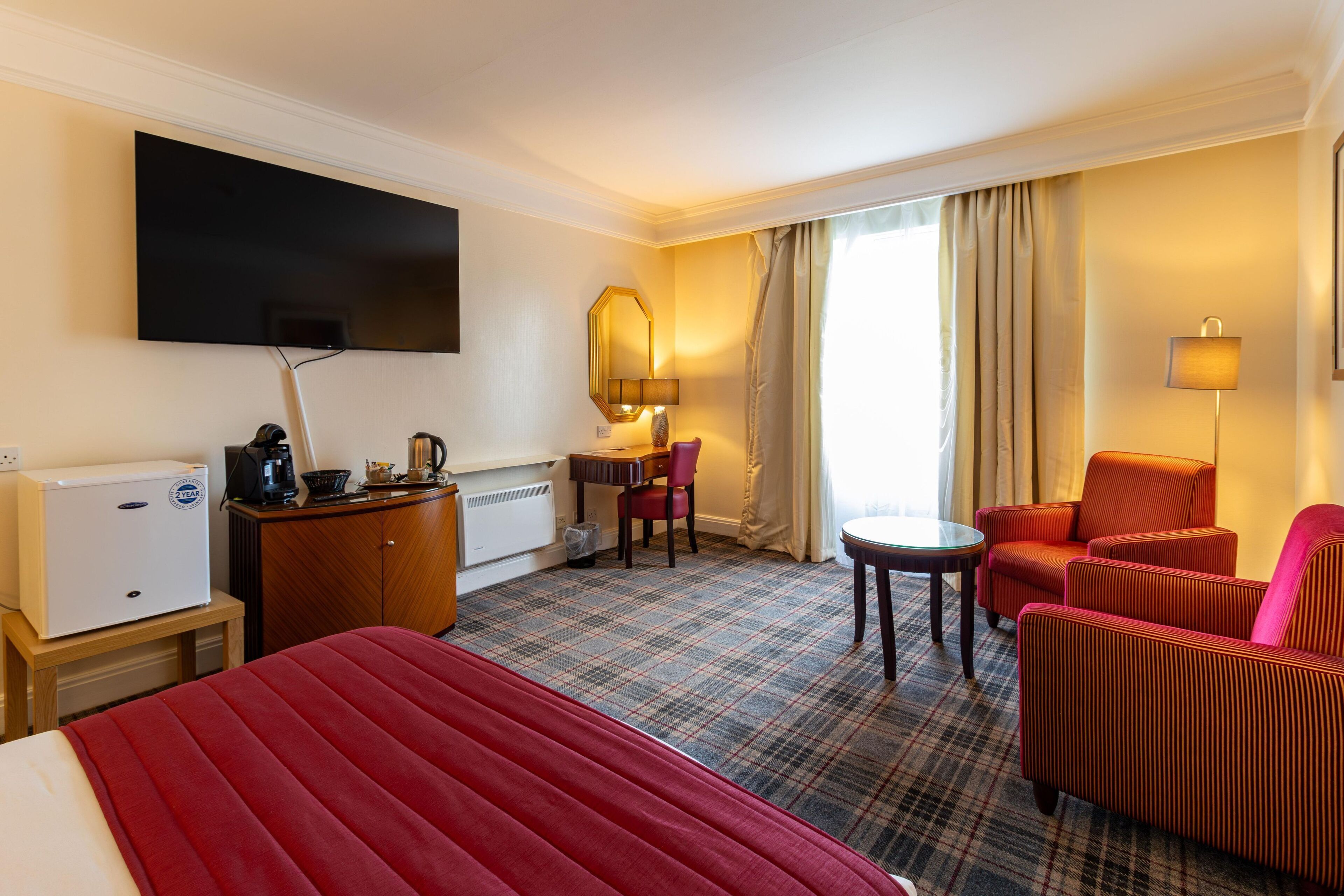 A hotel room with a bed, a desk, a chair, a sofa, a television, and a window with curtains.