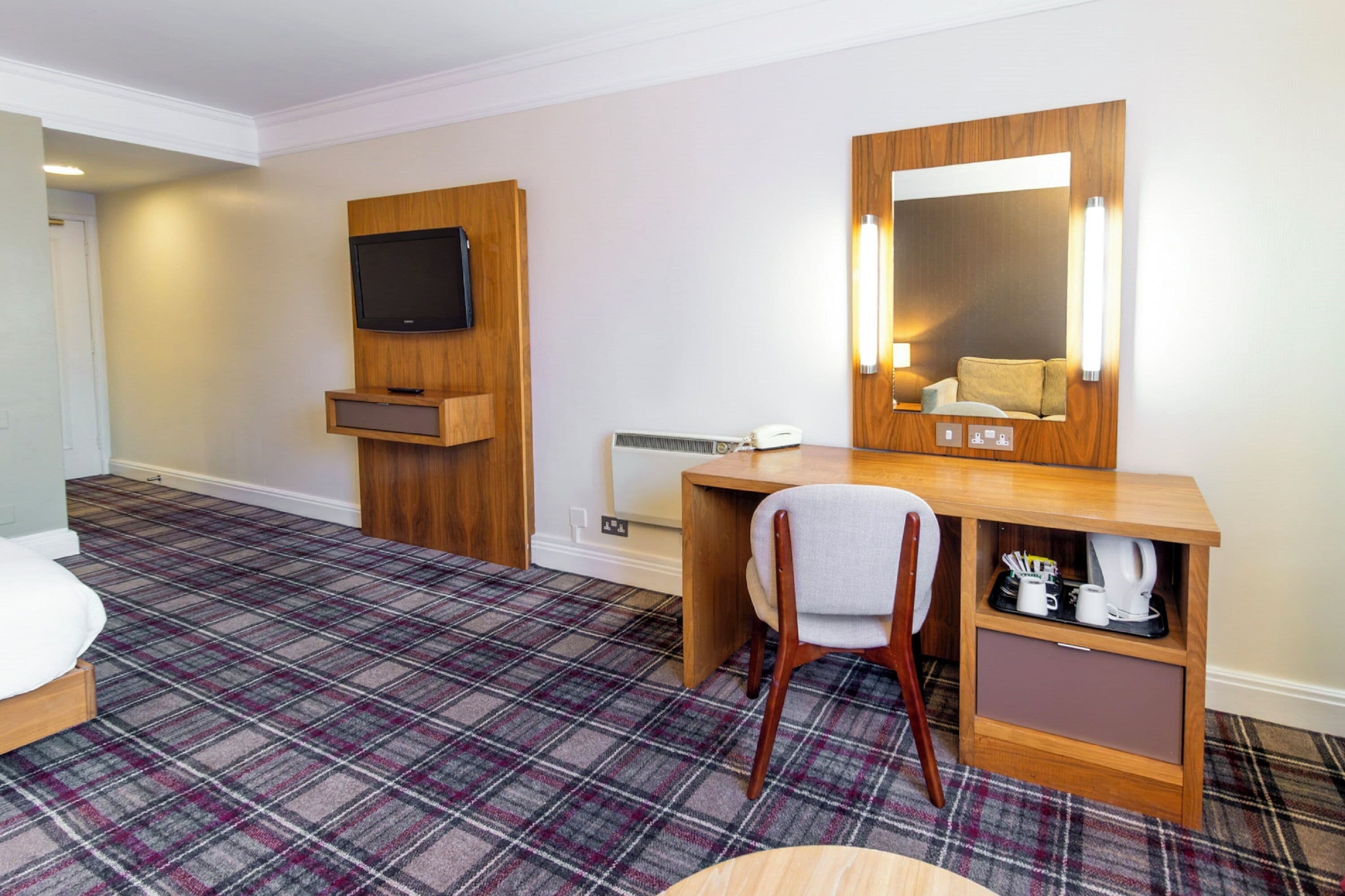 A hotel room with a plaid carpet, a bed, a desk with a chair, a TV mounted on the wall, and a mirror with lights.