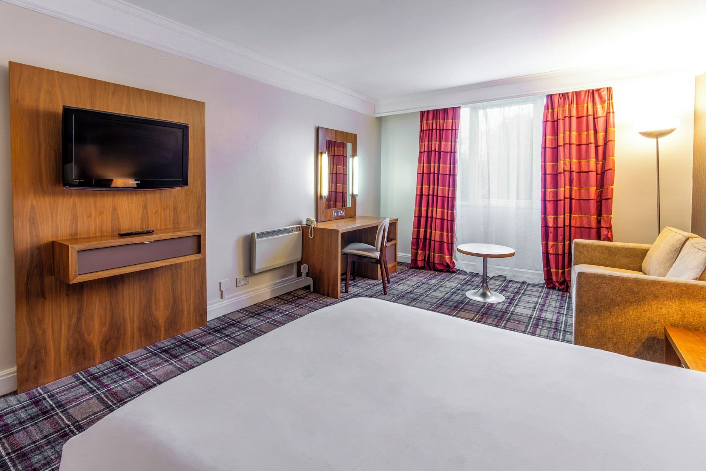 A hotel room with a large bed, a desk with a chair, a television mounted on the wall, and a window with red curtains.