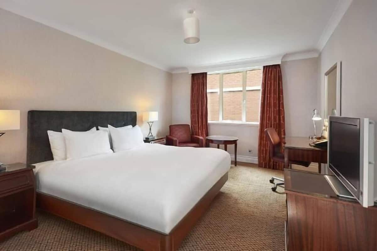 A hotel room with a large bed, a desk with a TV, a chair, a lamp, and a window with curtains.