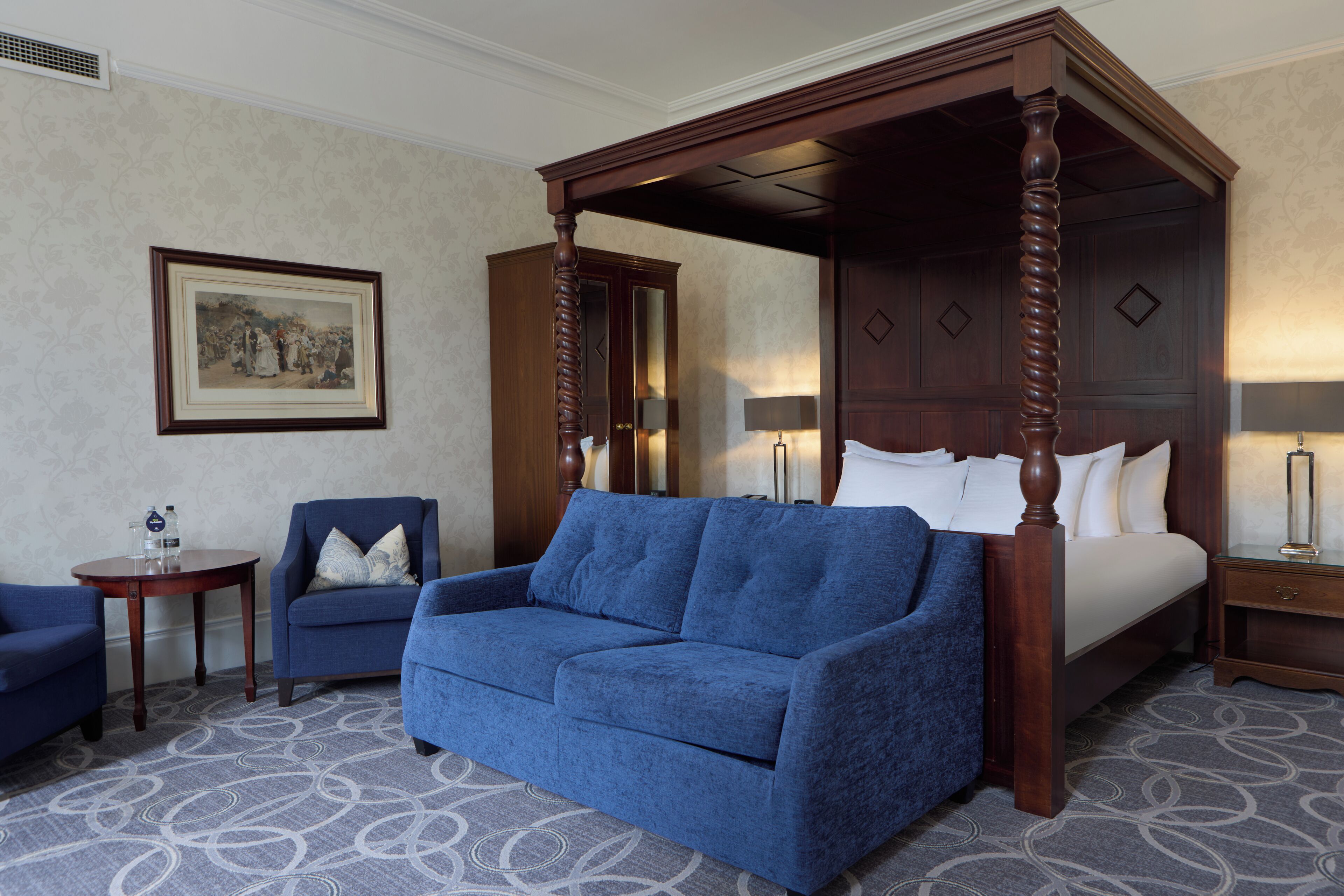 A four-poster bed with a wooden frame, a blue sofa, a small round table with a lamp, and a framed picture on the wall.
