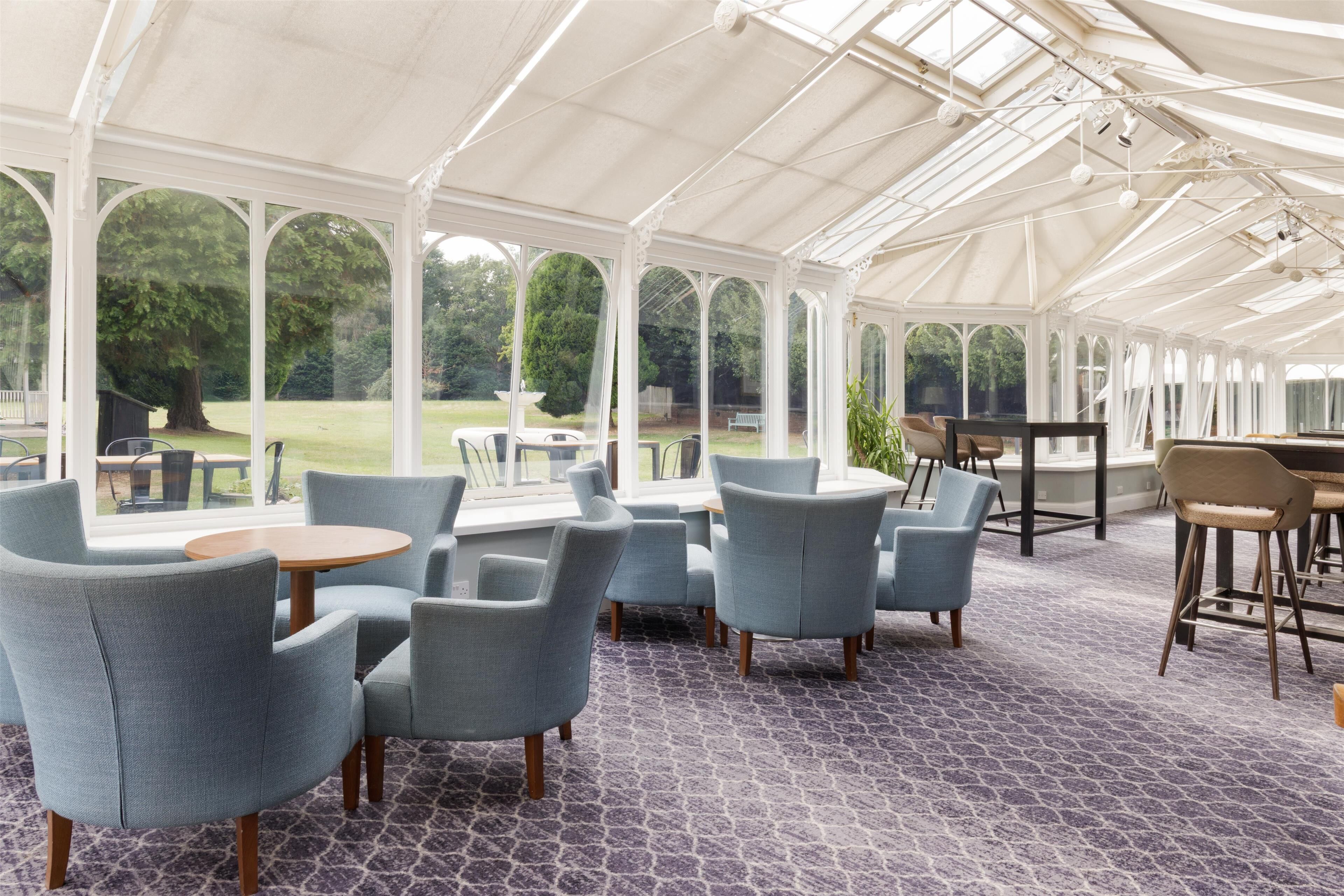 A conservatory with a patterned floor, seating area with round tables, and large windows offering a view of the outdoors.