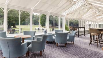A conservatory with a patterned floor, seating area with round tables, and large windows offering a view of the outdoors.