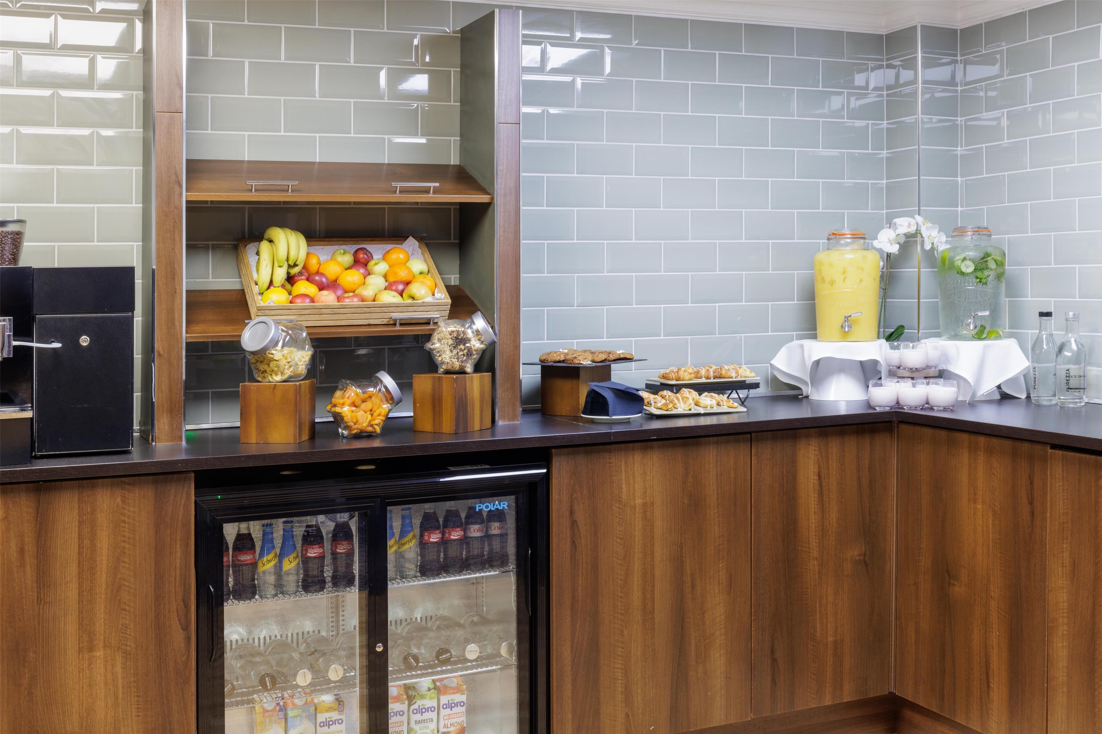 A breakfast bar with a fruit display, a refrigerator stocked with beverages, and a coffee machine.