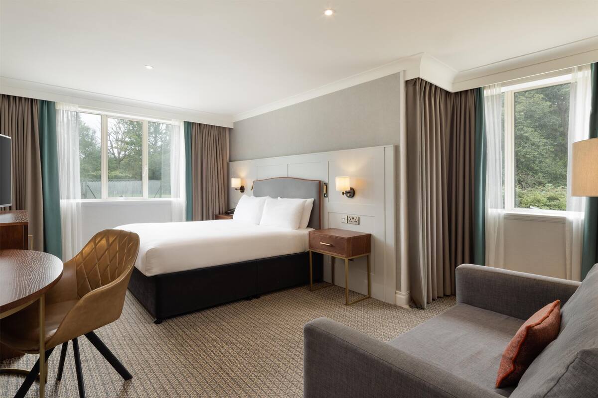 A hotel room with a bed, a desk, a chair, a sofa, and a window with curtains.