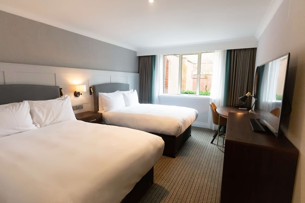 A hotel room with two beds, a desk, a TV, and a window with curtains.