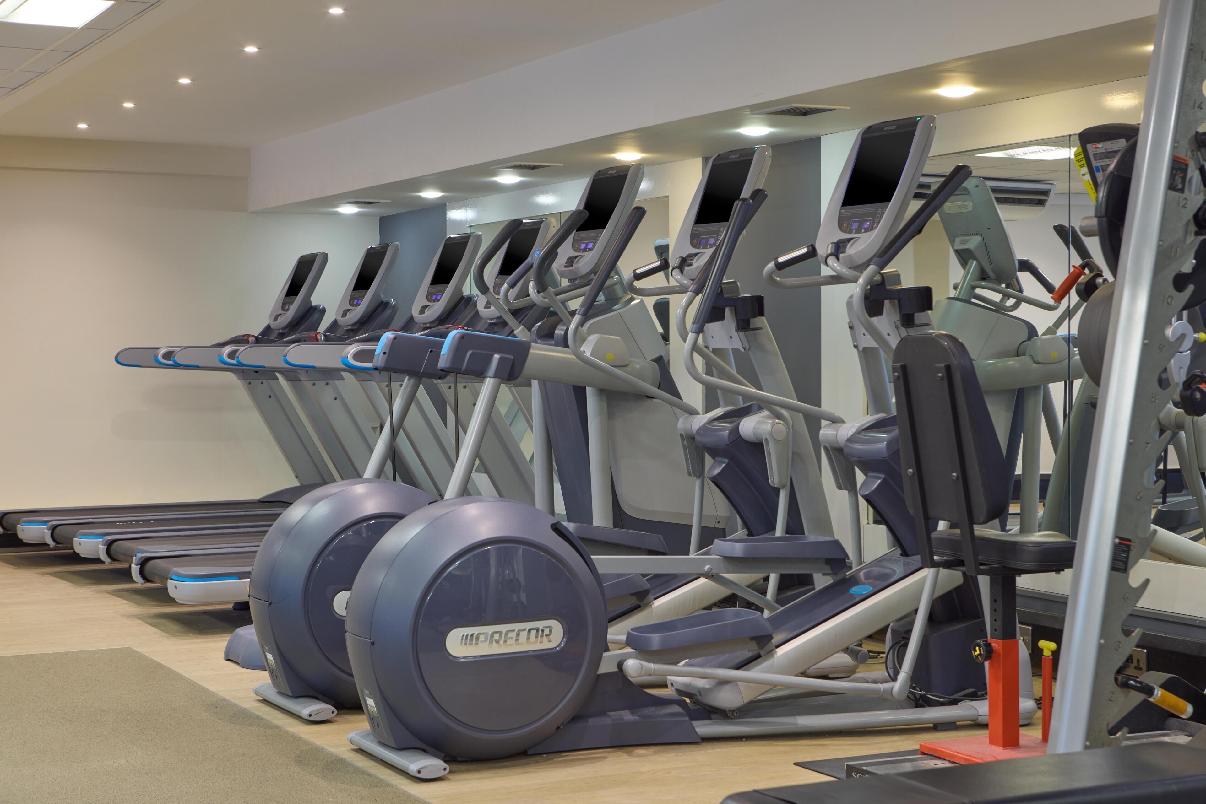 A row of treadmills in a gym with a mirrored wall and ceiling lights.