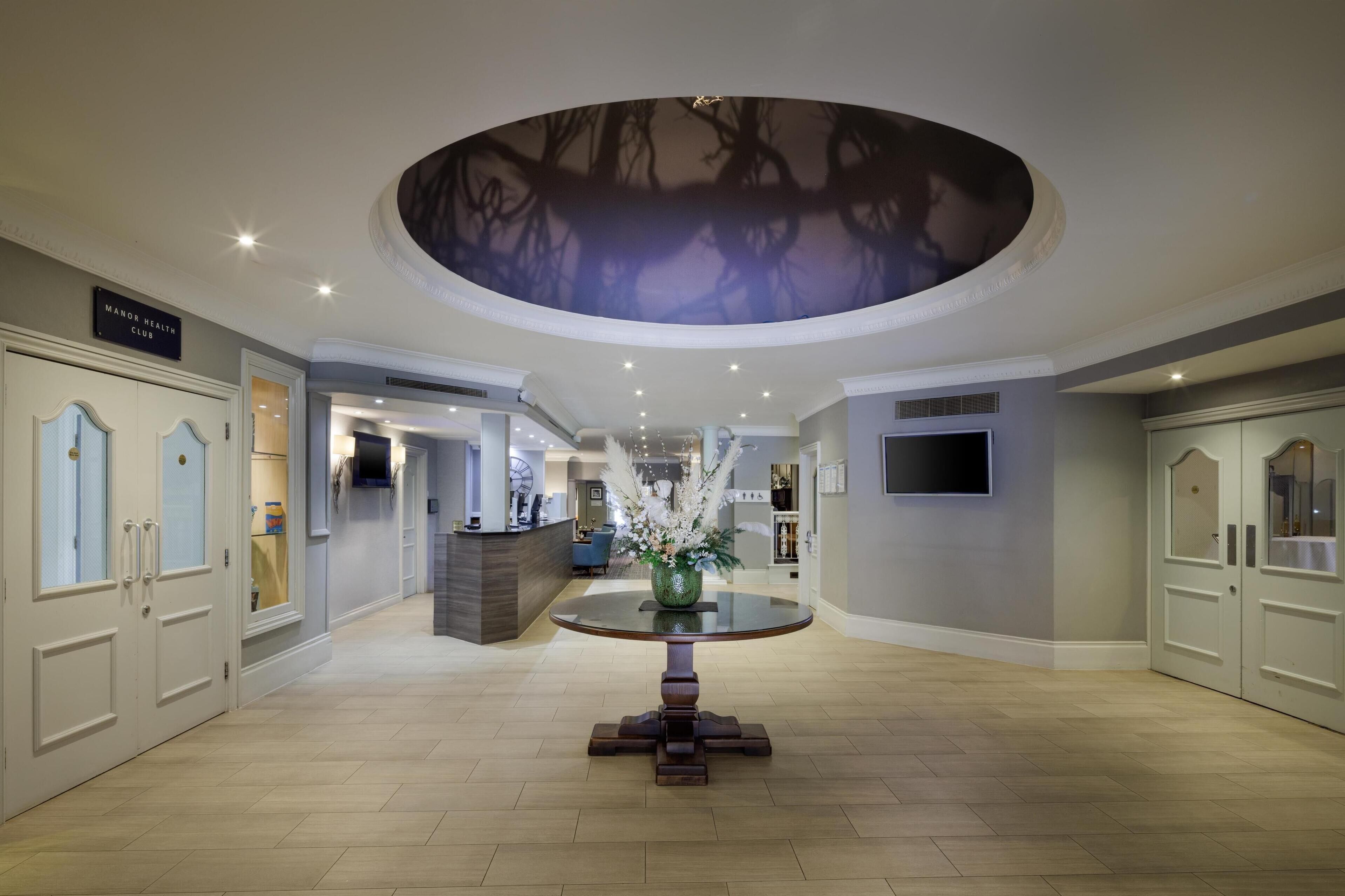 A spacious lobby with a round table, a ceiling mural, and a sign for the Manor Health Club.