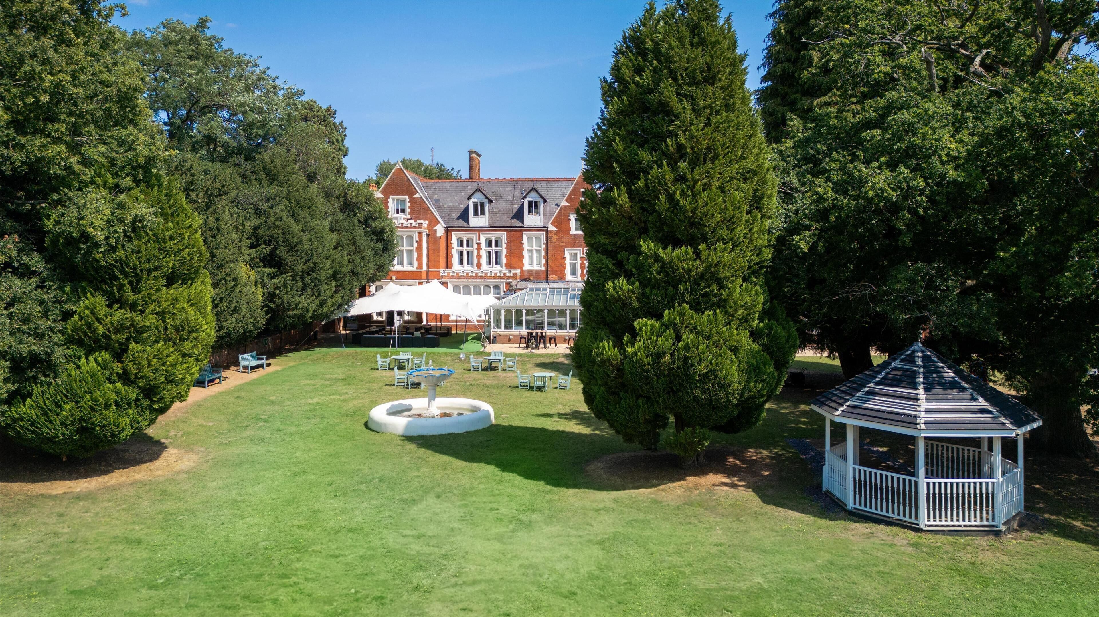 A large, traditional building with a symmetrical facade, a conservatory, and a well-maintained lawn with seating areas and a gazebo.