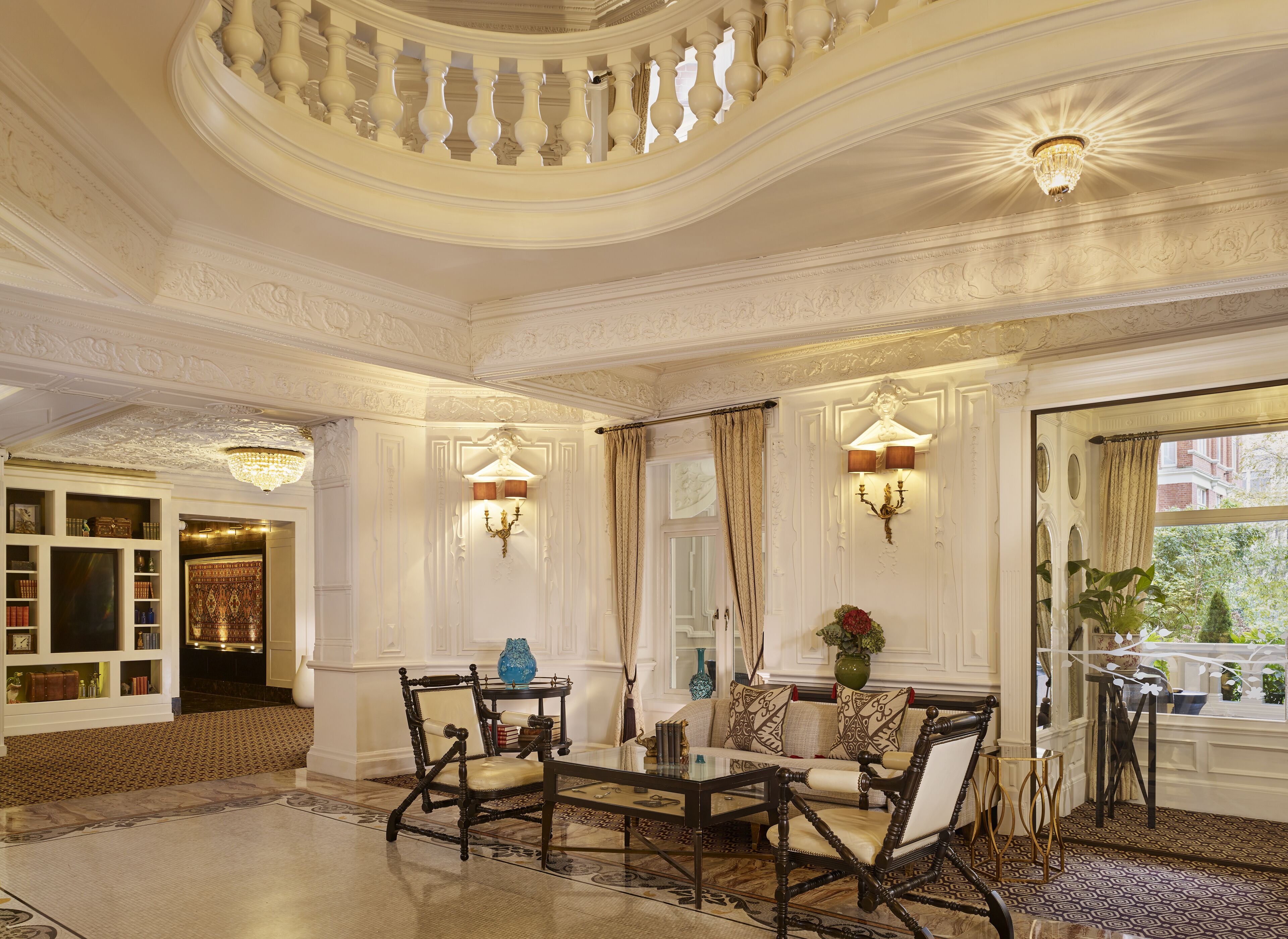 A grand room with ornate ceiling details, a large window with curtains, and a seating area with a table and chairs.