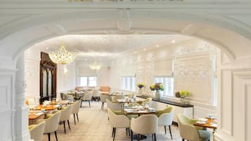 A spacious tea lounge with ornate white walls, chandeliers, and a variety of seating arrangements.