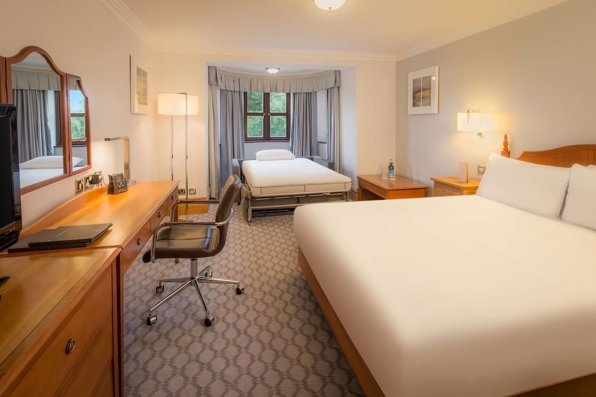 Een hotelkamer met een groot bed, een bureau met een stoel, een bureaulamp, een nachtkastje met een lamp en een raam met gordijnen.
