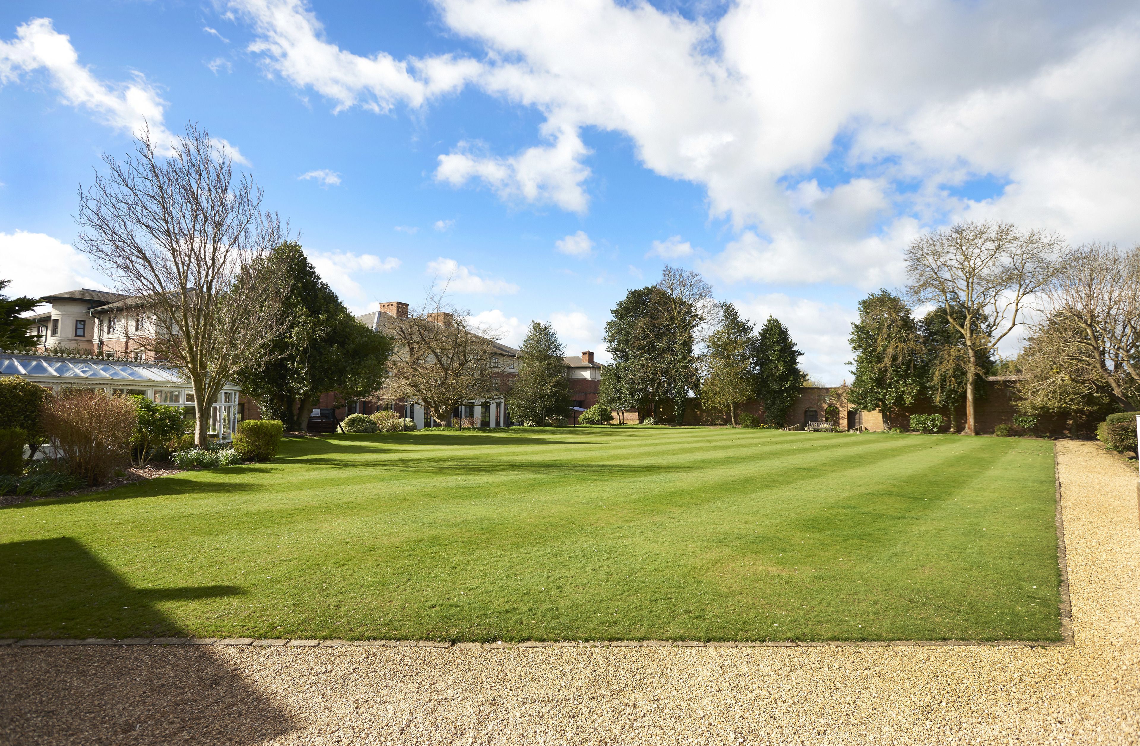 A well-maintained lawn with a gravel path, surrounded by mature trees and a few buildings in the background.
