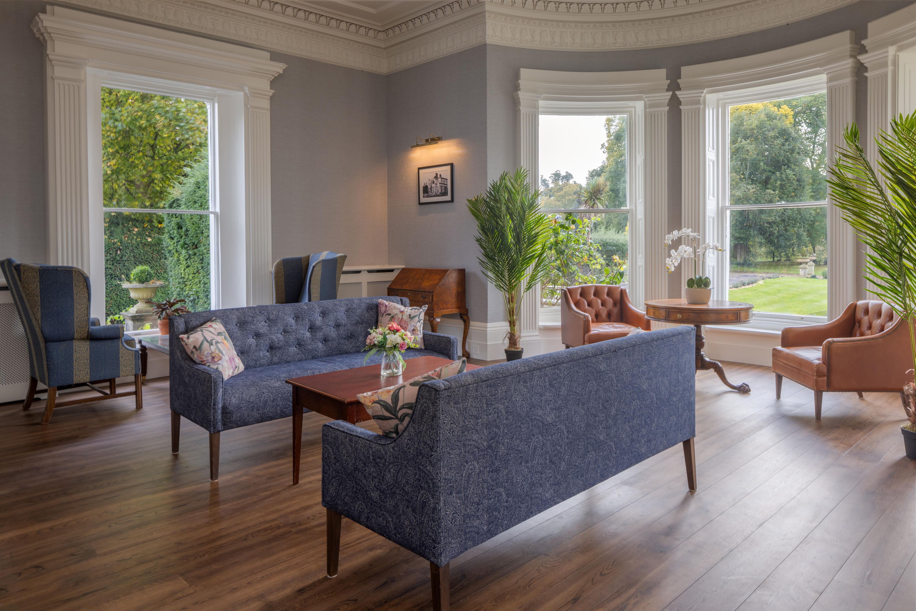 A spacious living room with a blue sofa, a brown leather armchair, and a wooden coffee table. There are large windows providing a view of the outdoors.
