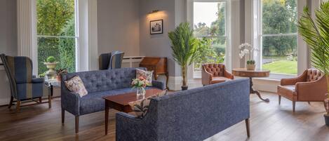 A spacious living room with a blue sofa, a brown leather armchair, and a wooden coffee table. There are large windows providing a view of the outdoors.