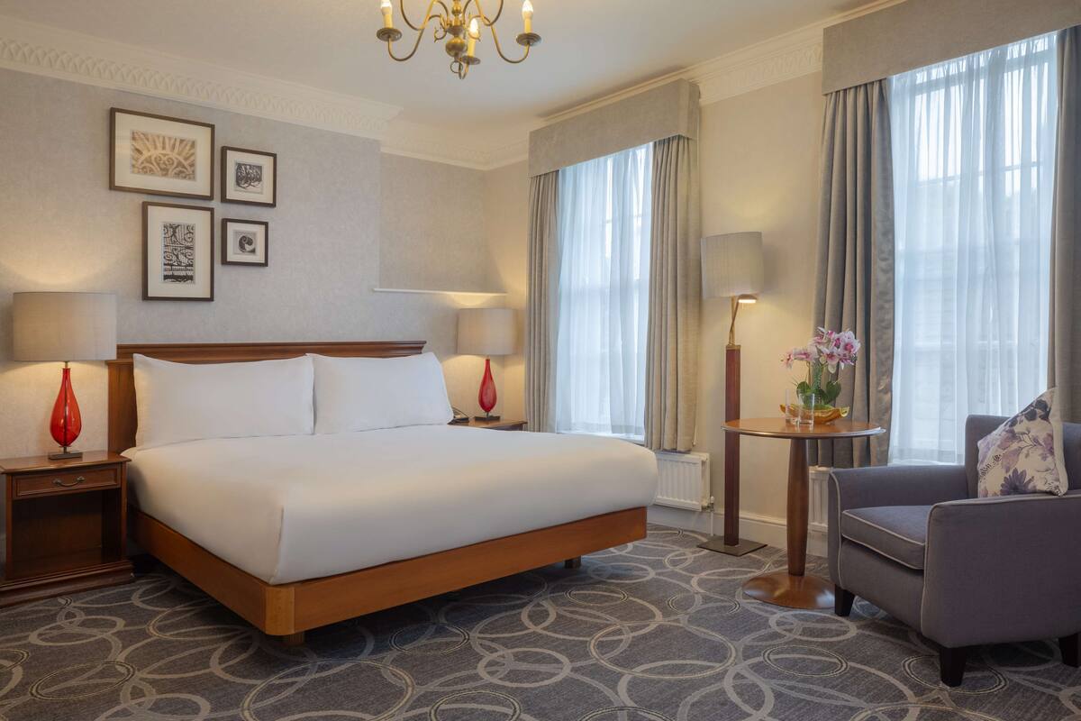 Een hotelkamer met een groot bed, twee nachtkastjes met lampen, een stoel, een klein tafeltje met een vaas en een kroonluchter.