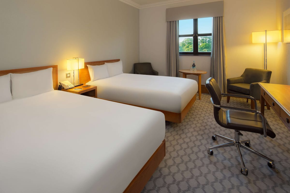 Een hotelkamer met twee bedden, een bureau, een stoel, een lamp en een raam met uitzicht op bomen.