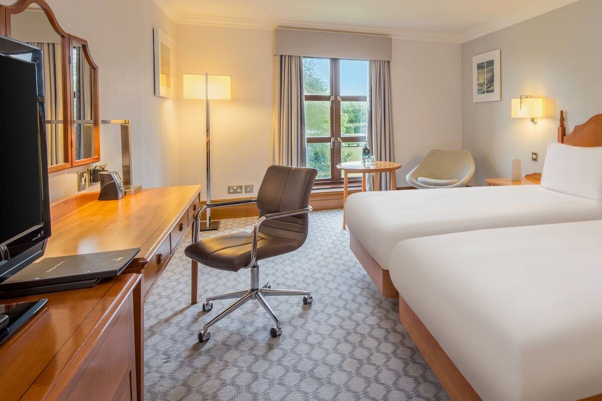 Een hotelkamer met twee bedden, een bureau met een laptop, een stoel, een televisie en een raam met gordijnen.