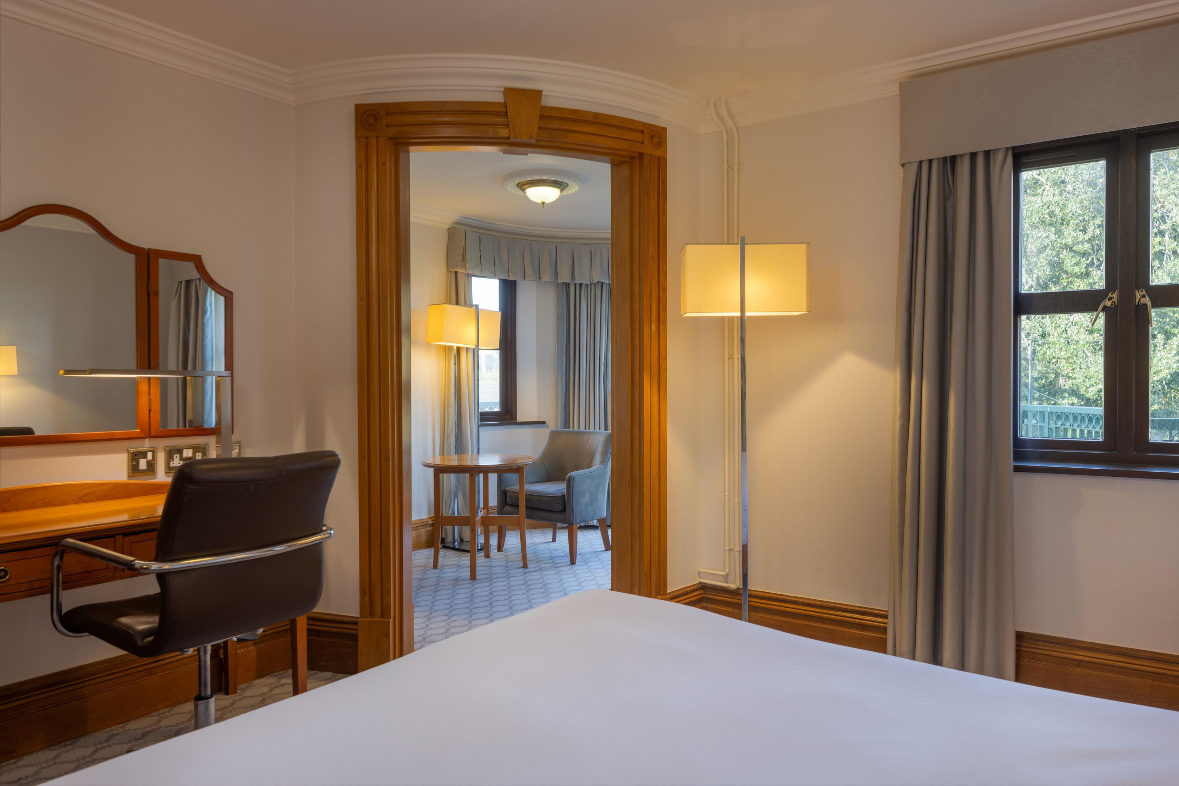 A hotel room with a bed, a desk with a chair, a mirror, a lamp, and a view of the outdoors through a window.