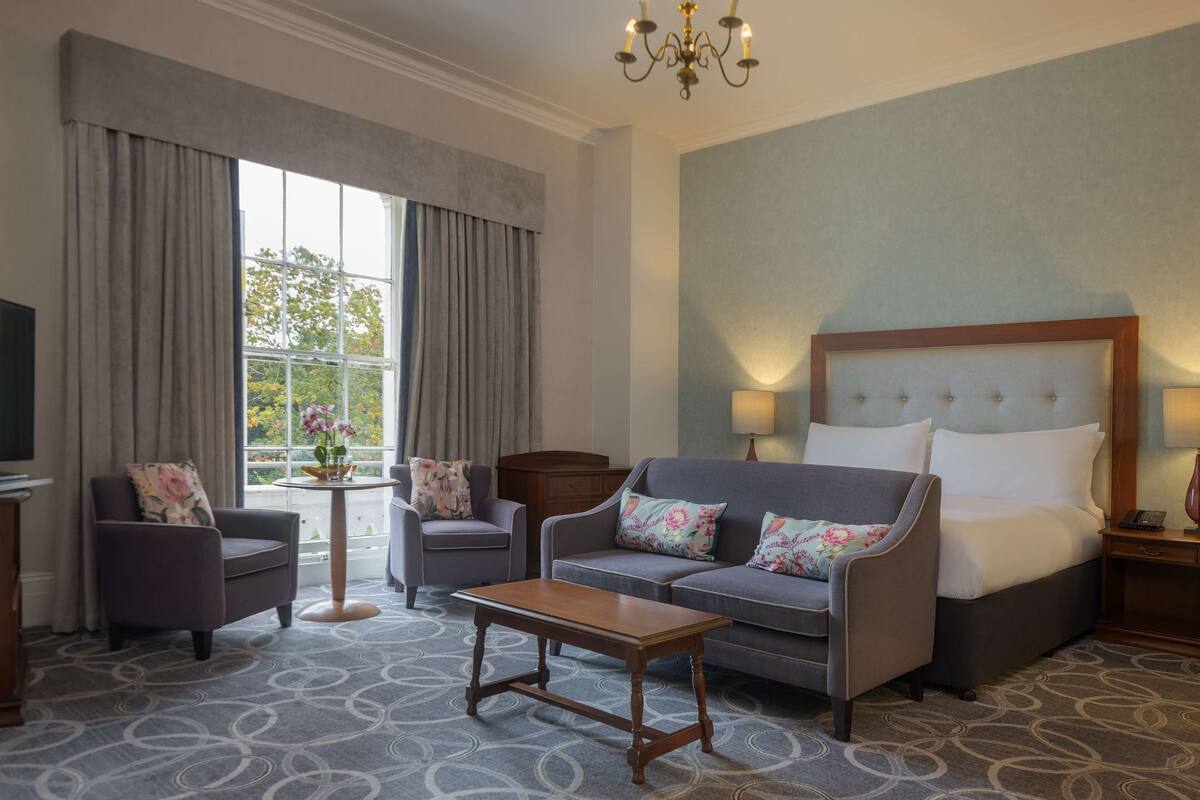 Een hotelkamer met een bank, twee fauteuils, een bijzettafeltje, een bed met hoofdeinde, een nachtkastje, een lamp en een televisie.