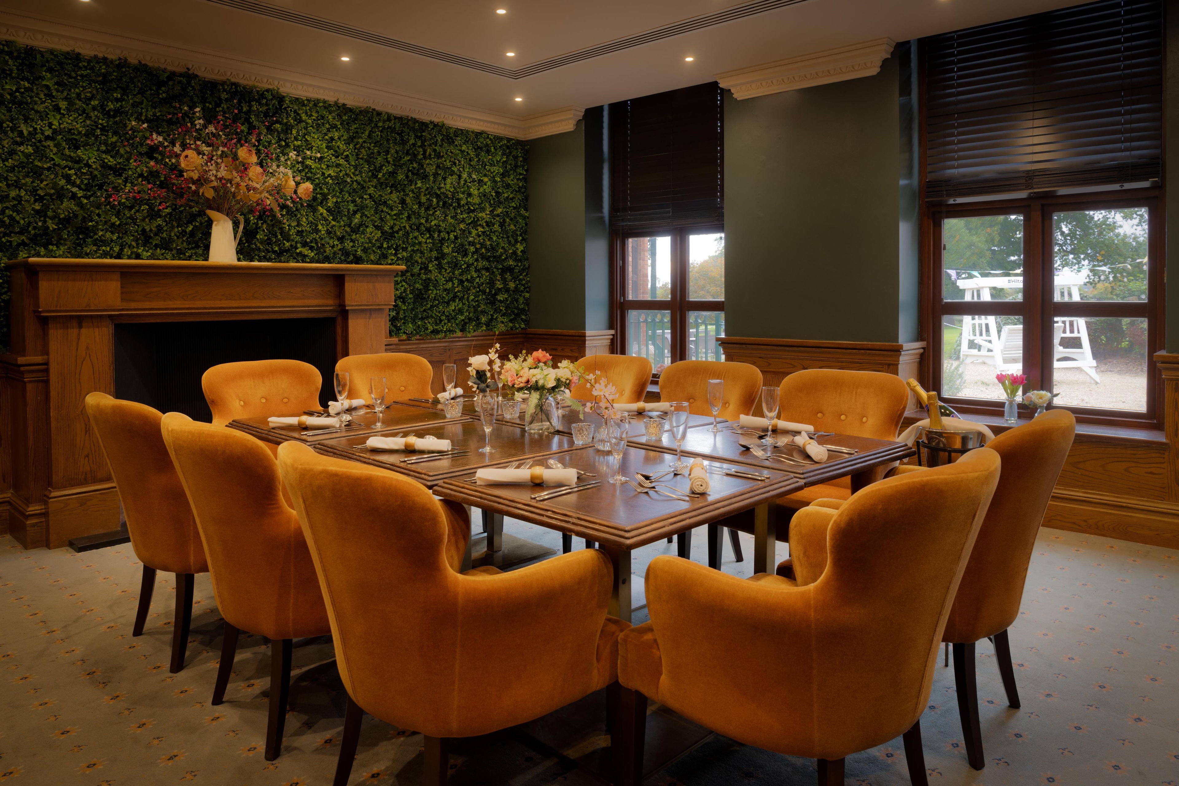 A dining room with a long table set for eight, surrounded by orange chairs, a green wall with foliage, and a fireplace.