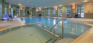 Une piscine intérieure à l’eau claire, entourée de murs dotés de grandes fenêtres, et un fauteuil de relaxation sur la gauche.