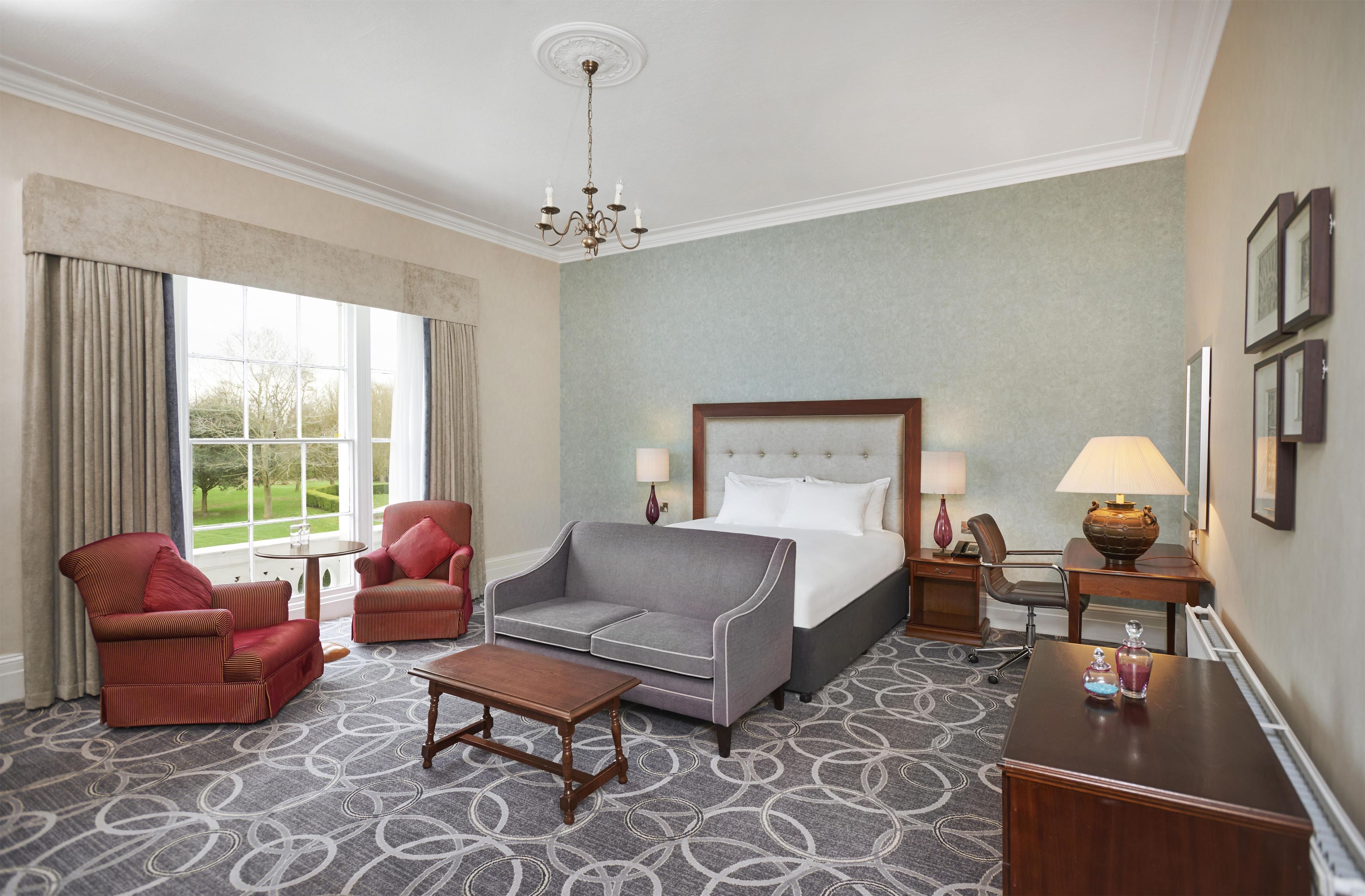 Une chambre d’hôtel comprenant un lit, deux fauteuils, une table basse, un bureau avec une lampe et une vue sur l’extérieur.