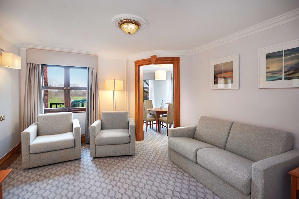 Hotelkamer met een bank, twee fauteuils, een eettafel met stoelen, uitzicht door een raam en een deur naar een andere kamer.