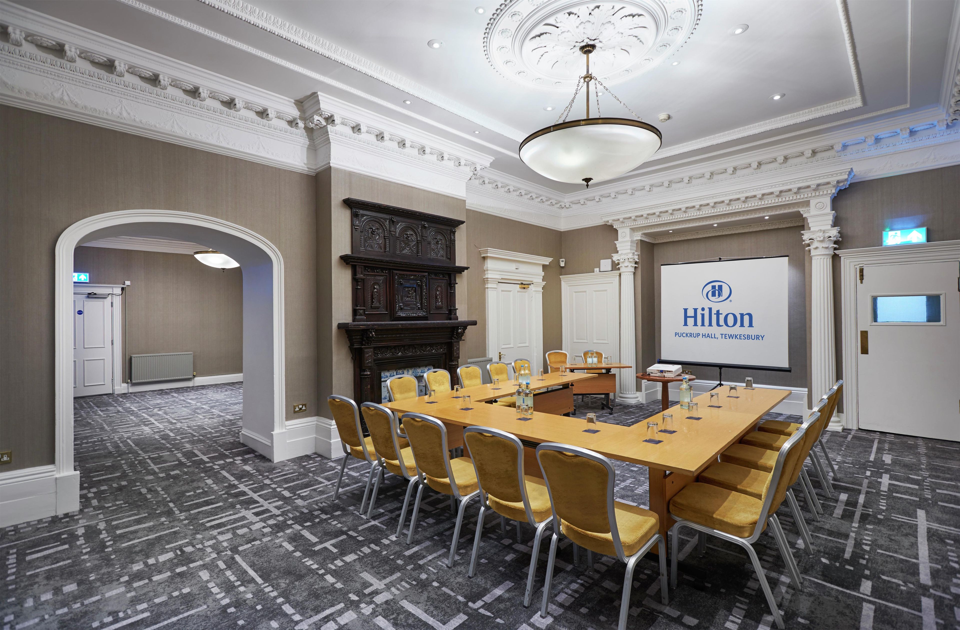 Un salón de conferencias con una larga mesa, sillas amarillas, una chimenea y una pantalla grande que muestra el logotipo de Hilton.