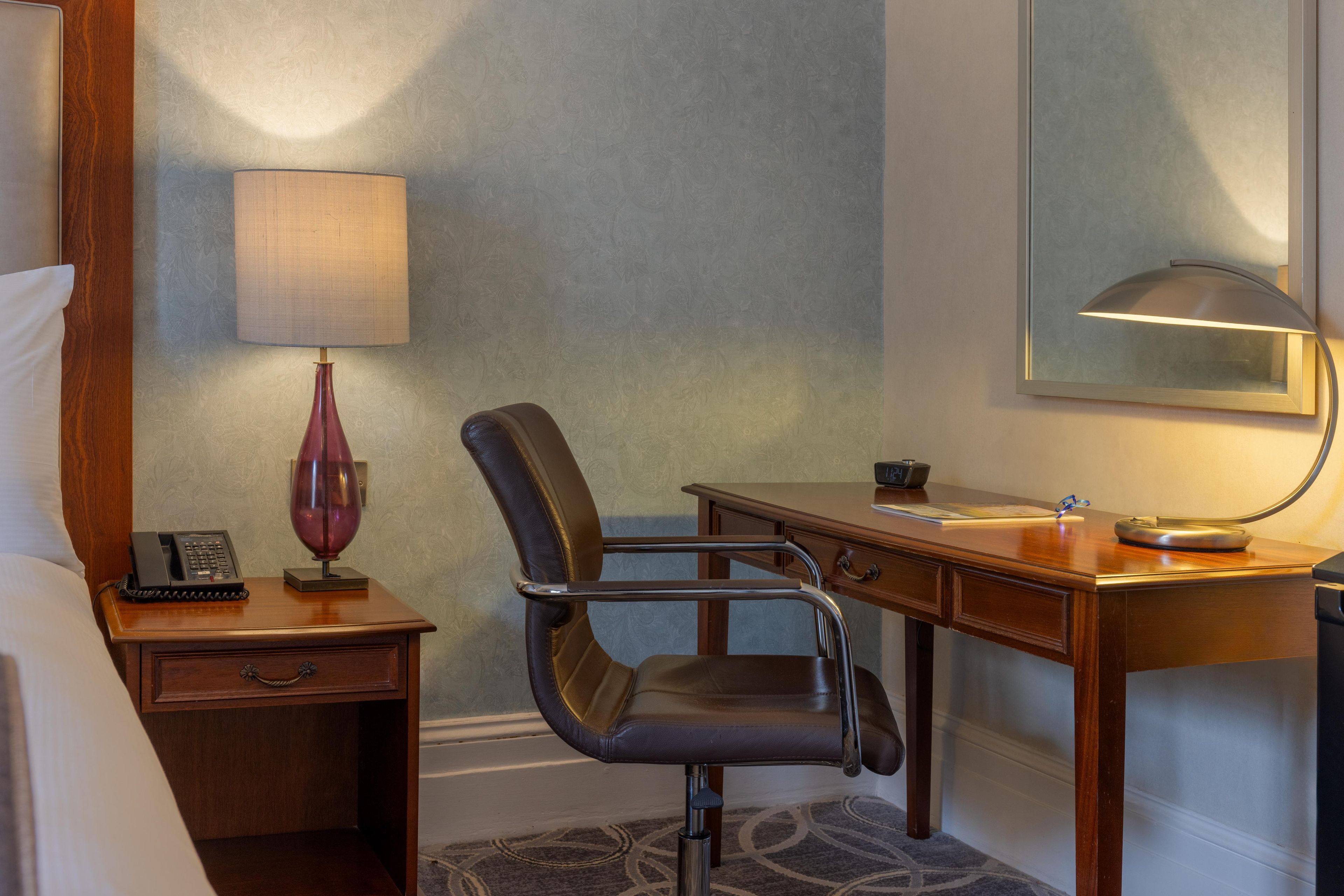 Ein Hotelzimmer mit einem Bett, einem Schreibtisch, einem Stuhl, einer Lampe, einem Telefon und einem Spiegel.