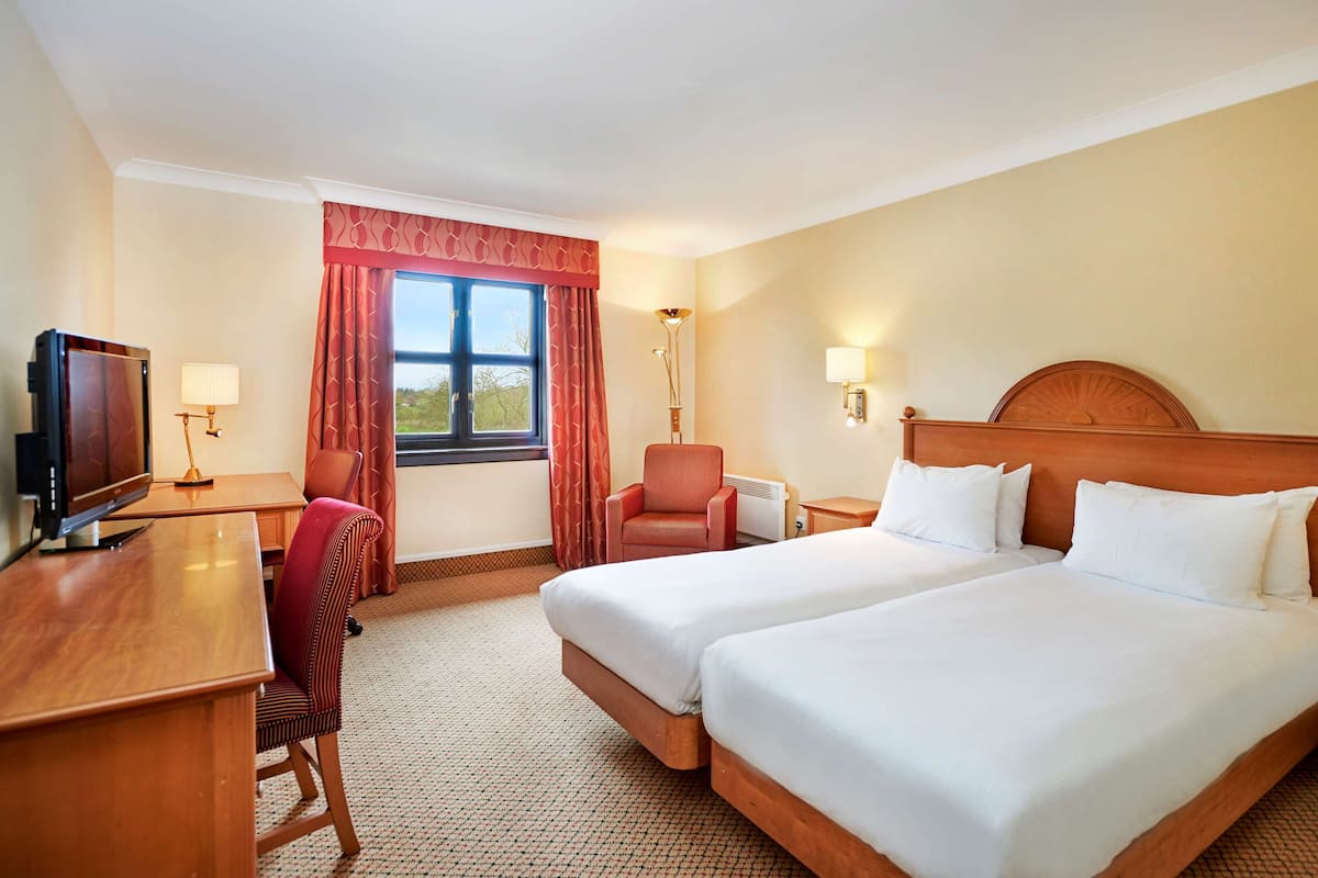 Een hotelkamer met twee bedden, een bureau met een televisie, een stoel, een lamp en een raam met gordijnen.