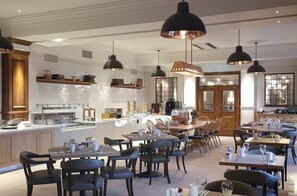 Un espace de restauration moderne avec plusieurs tables et chaises, une cuisine en arrière-plan et des luminaires suspendus.