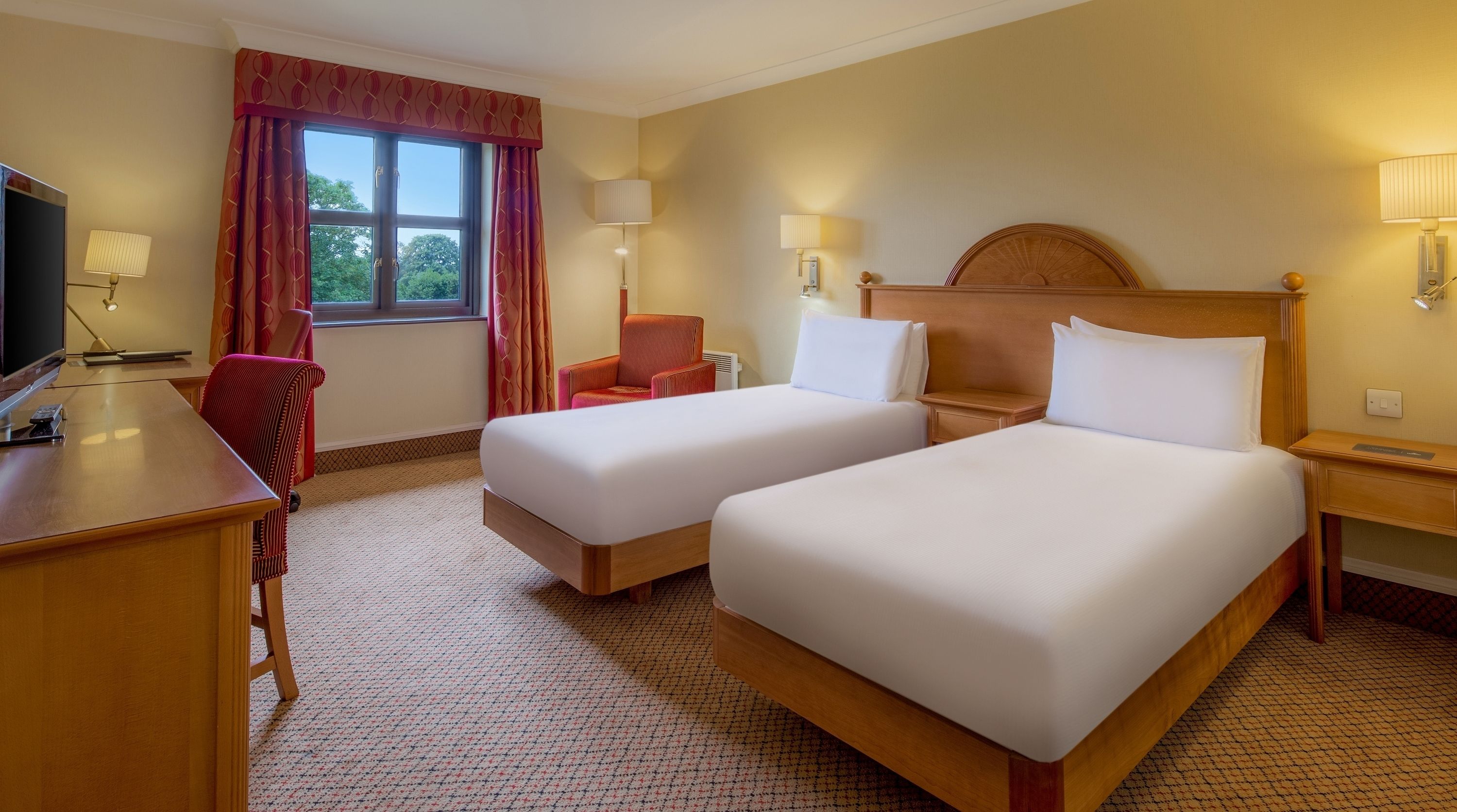 A hotel room with two single beds, a desk with a computer, a chair, and a window with curtains.