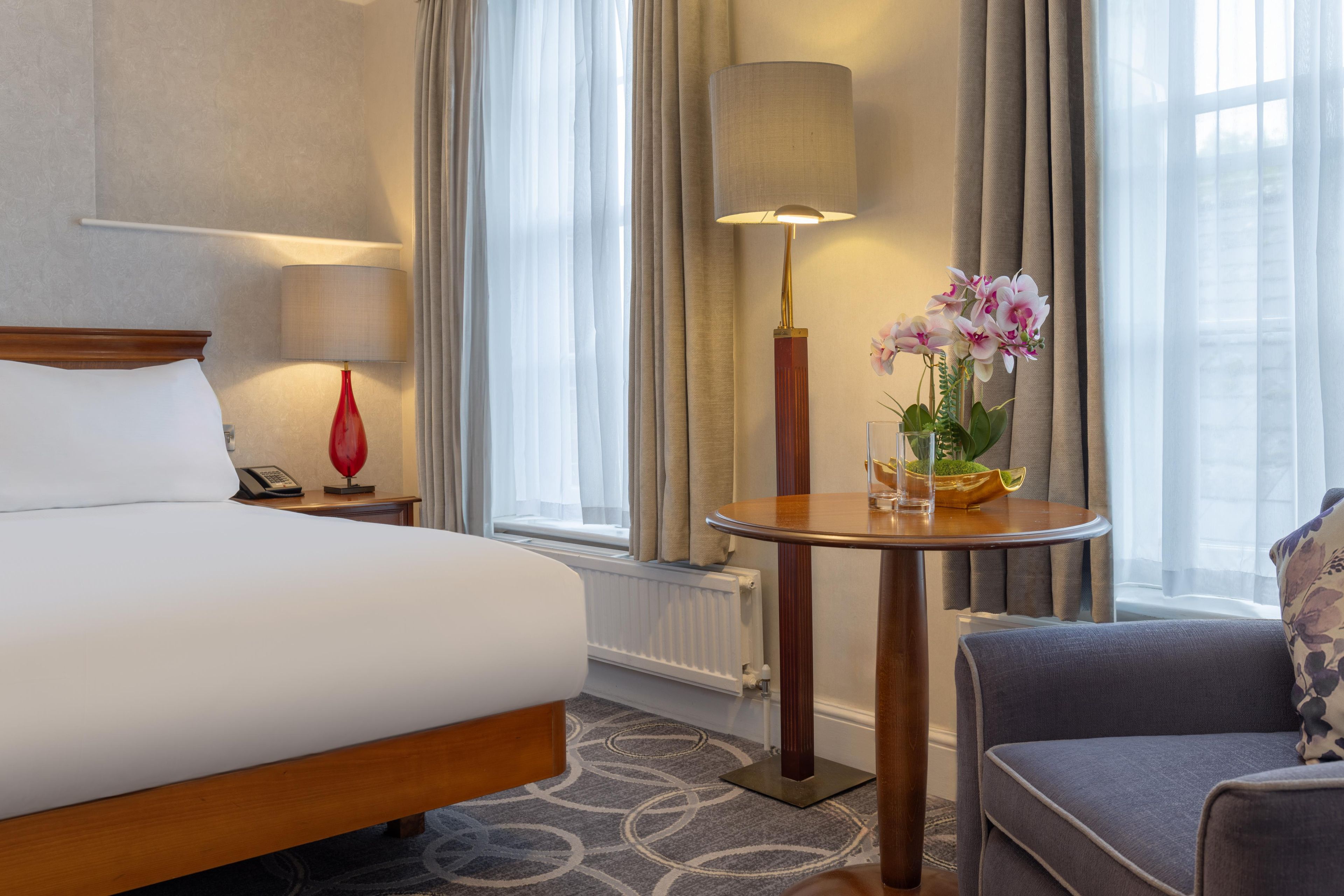 Una habitación de hotel con cama, mesita de noche, lámpara, silla y una ventana con cortinas.