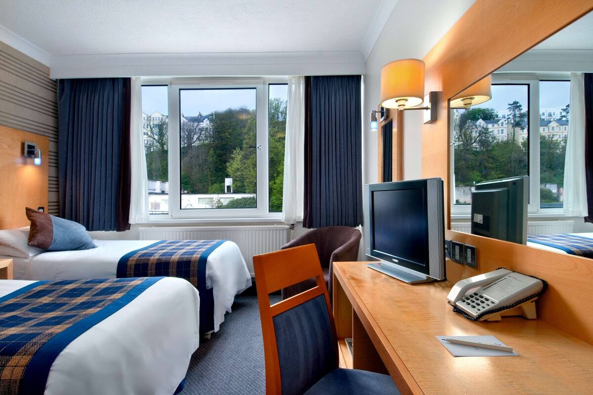 A hotel room with two beds, a desk with a computer and phone, a chair, and large windows offering a view of trees and buildings.