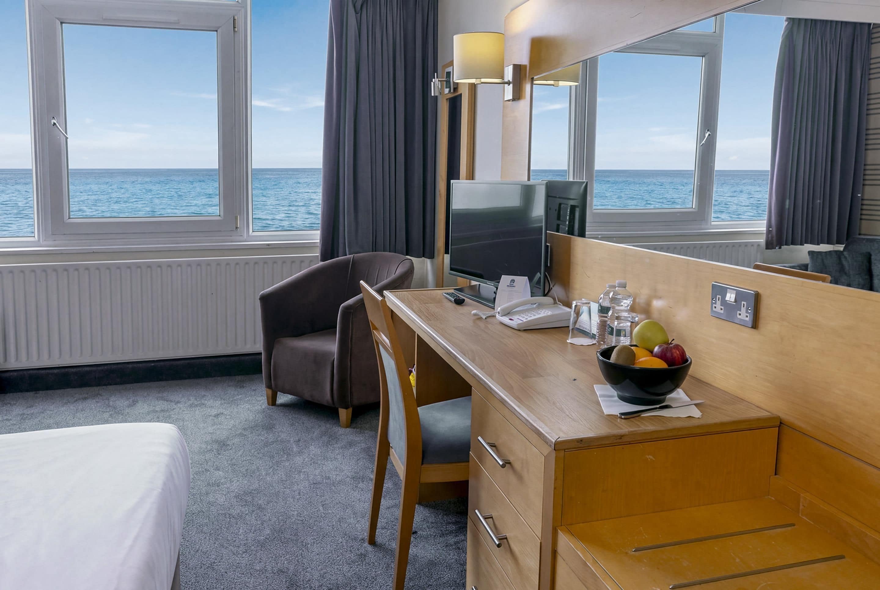 Hotellihuone, jossa on sänky, työpöytä, tuoli, televisio, puhelin, hedelmäkulho ja näkymä merelle.